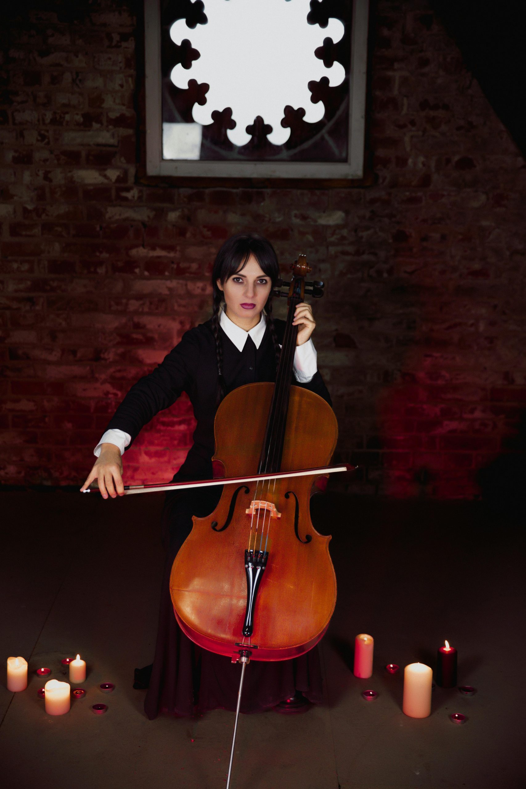 A woman dressed in Gothic attire plays a cello surrounded by candles, creating a moody ambiance.