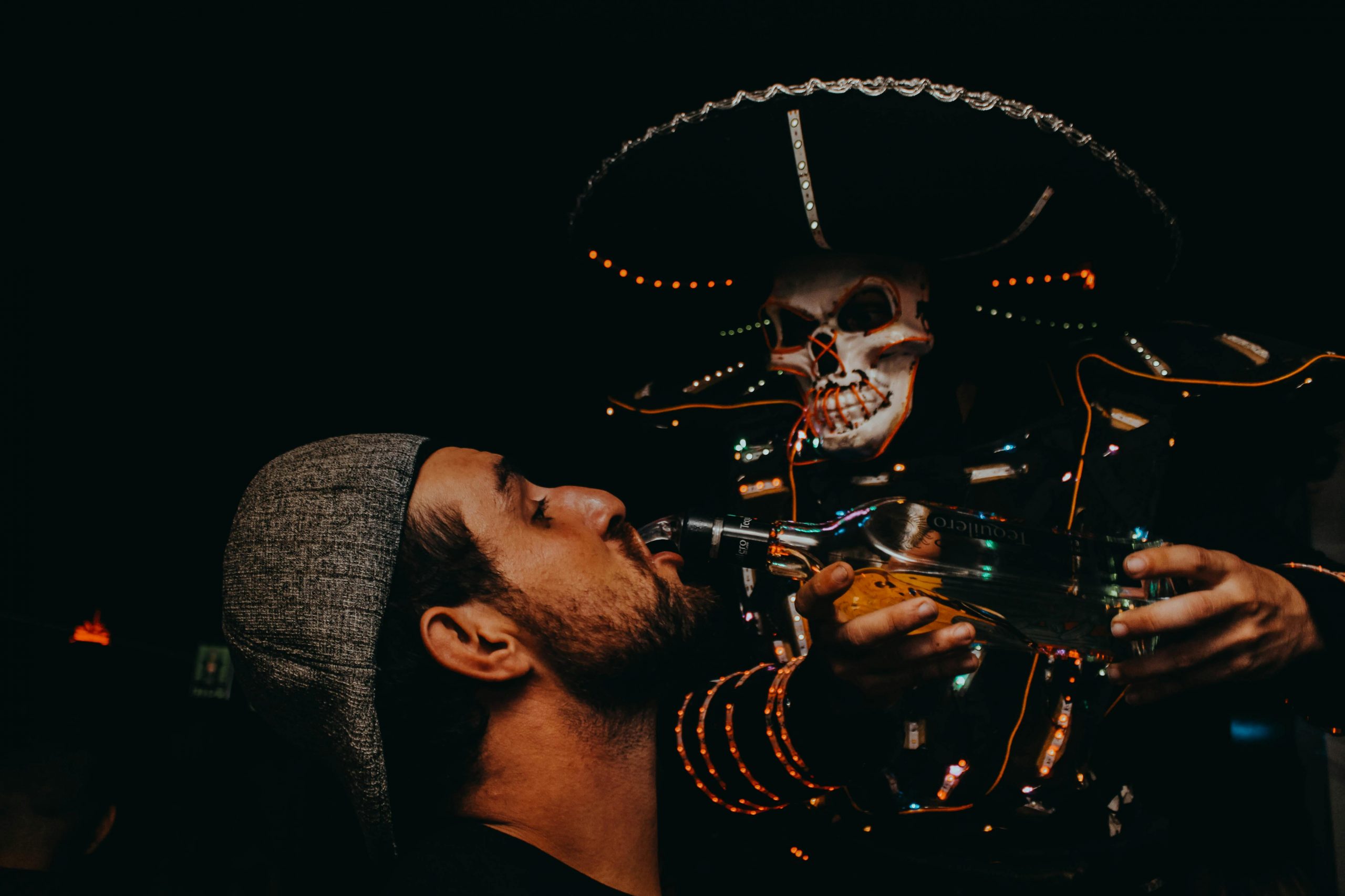 A festive night scene featuring a man enjoying a drink served by a vibrant skull-masked performer.
