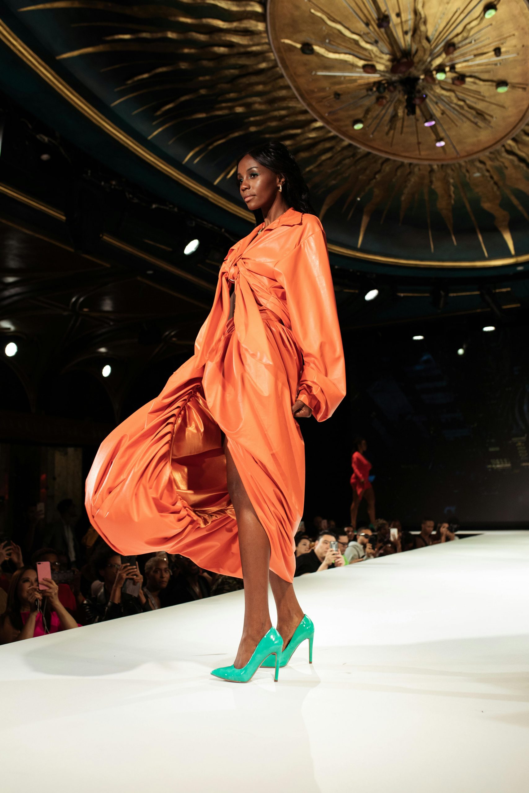 An African American model struts confidently in an orange dress during a fashion show.